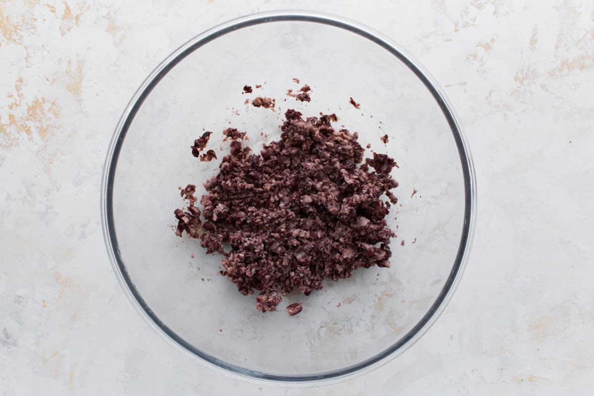 A clear mixing bowl with roughly mashed black beans, the first step in preparing black bean burger mixture.