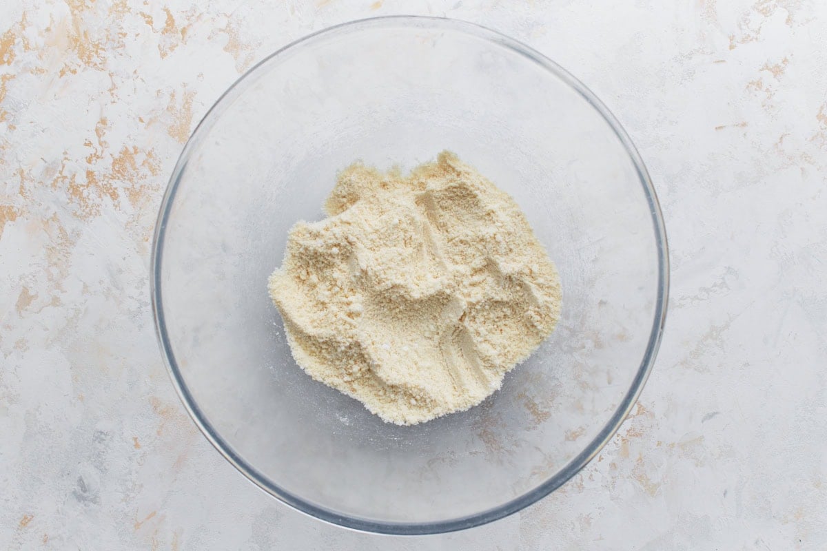 A glass mixing bowl filled with almond flour on a light, textured surface.