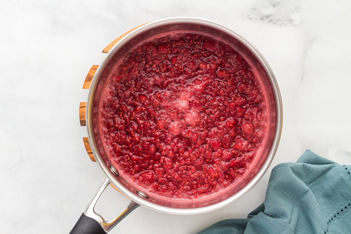 Overhead view of a saucepan filled with simmering raspberries, sugar, lemon juice, and water, starting to break down and release juices.