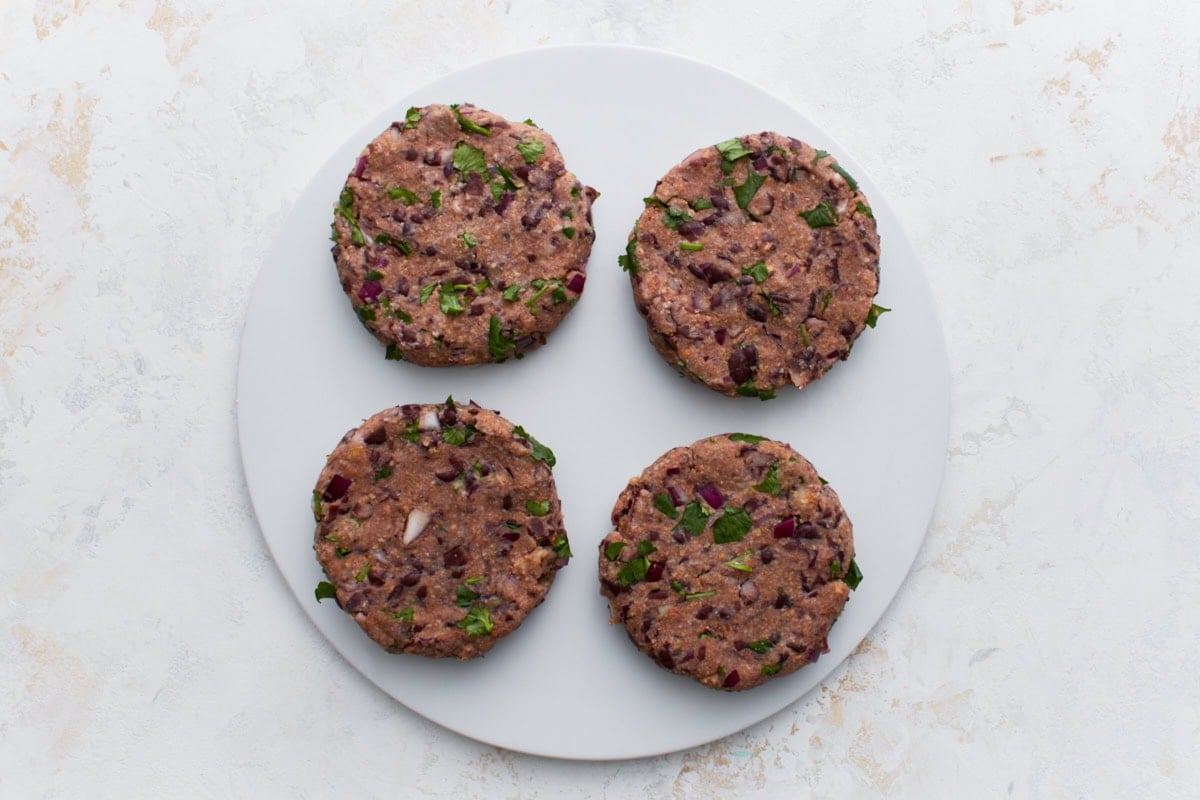 Four uncooked black bean burger patties shaped and arranged on a white plate, ready to be cooked.