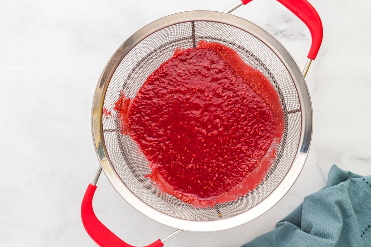 Straining raspberry puree through a fine mesh sieve, separating the smooth sauce from the seeds over a white countertop.