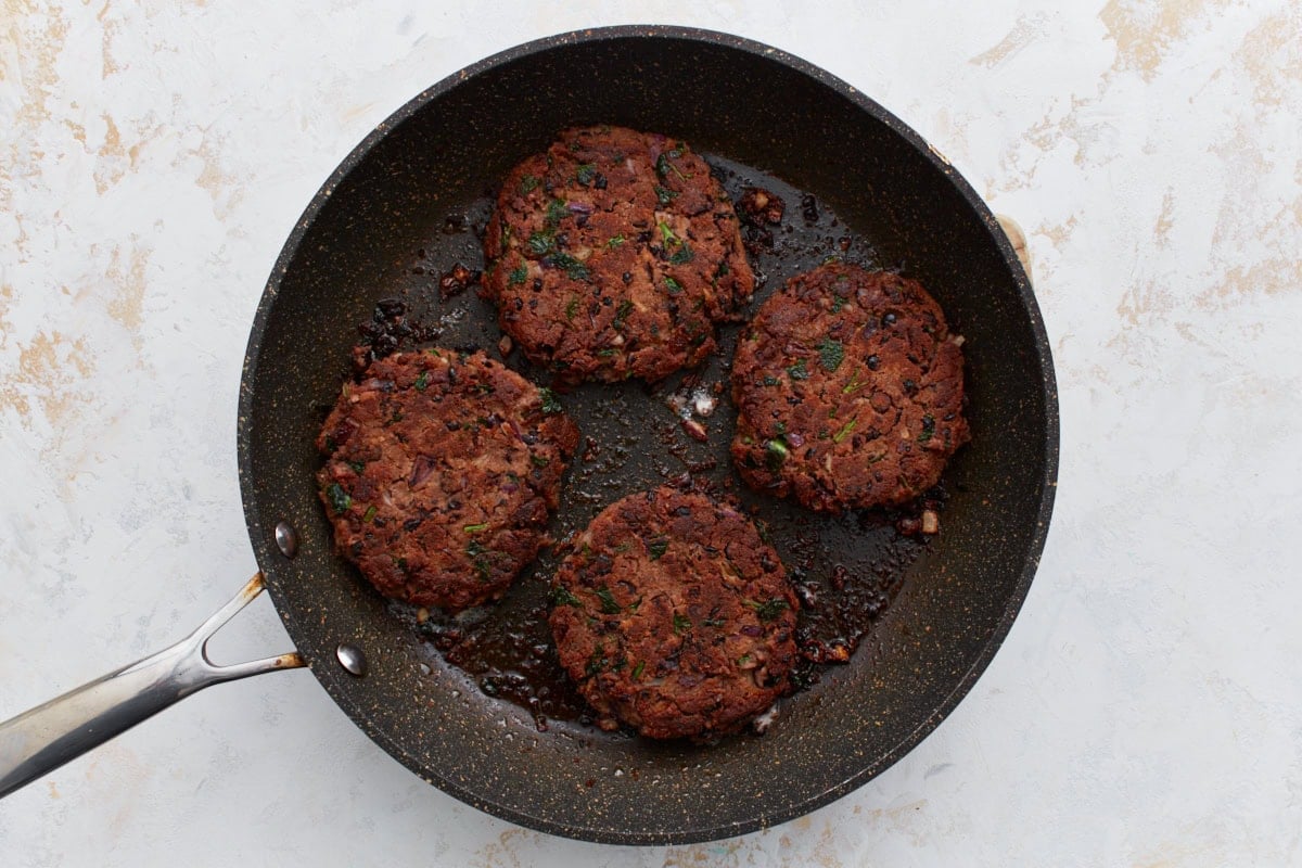 Four black bean burger patties cooking in a skillet, searing to a crispy golden-brown finish.