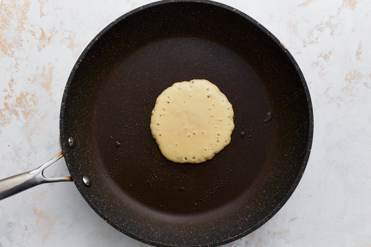 A single pancake cooking in a non-stick skillet, with bubbles beginning to form on the surface.