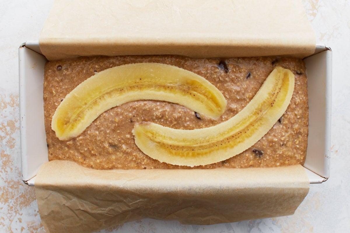 A parchment-lined loaf pan filled with banana bread batter, topped with two long banana slices.