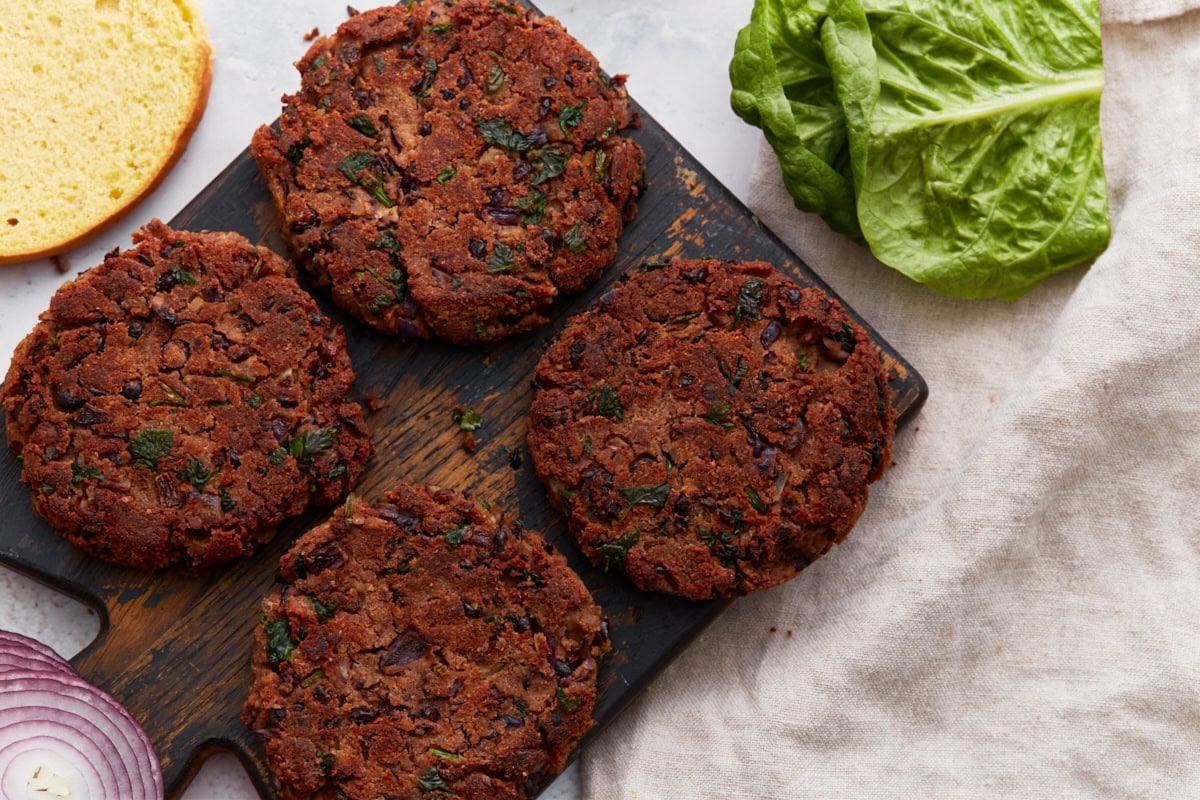 A top-down view of cooked black bean burgers on a dark wooden board with fresh lettuce, red onion, and a gluten-free bun nearby.