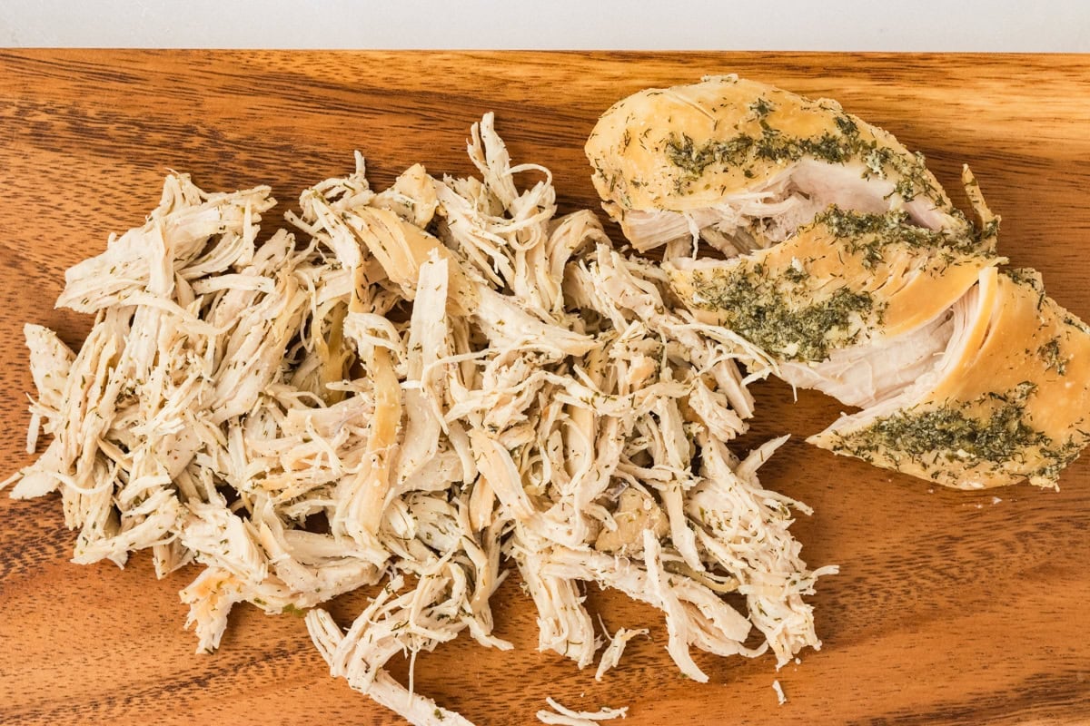 Shredded cooked chicken breast on a wooden cutting board, seasoned with herbs.