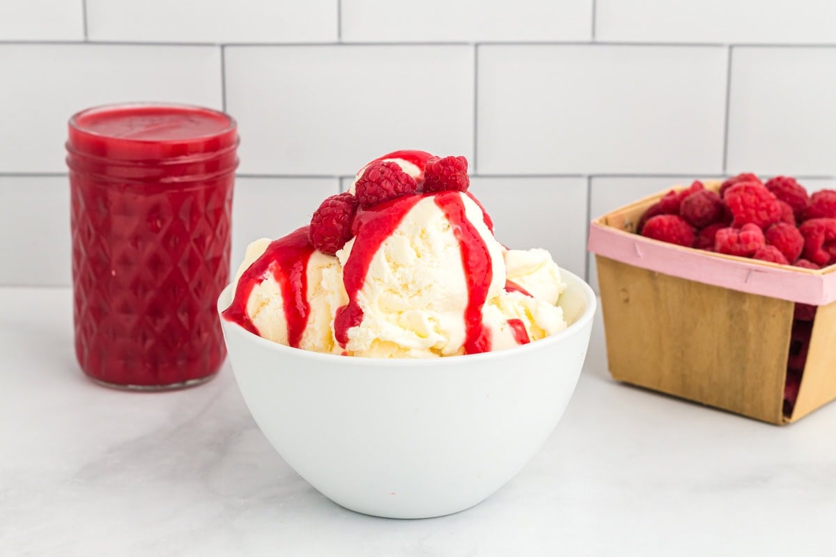 Vanilla ice cream in a white bowl topped with raspberry coulis and fresh raspberries, with a mason jar of coulis and a basket of berries in the background.