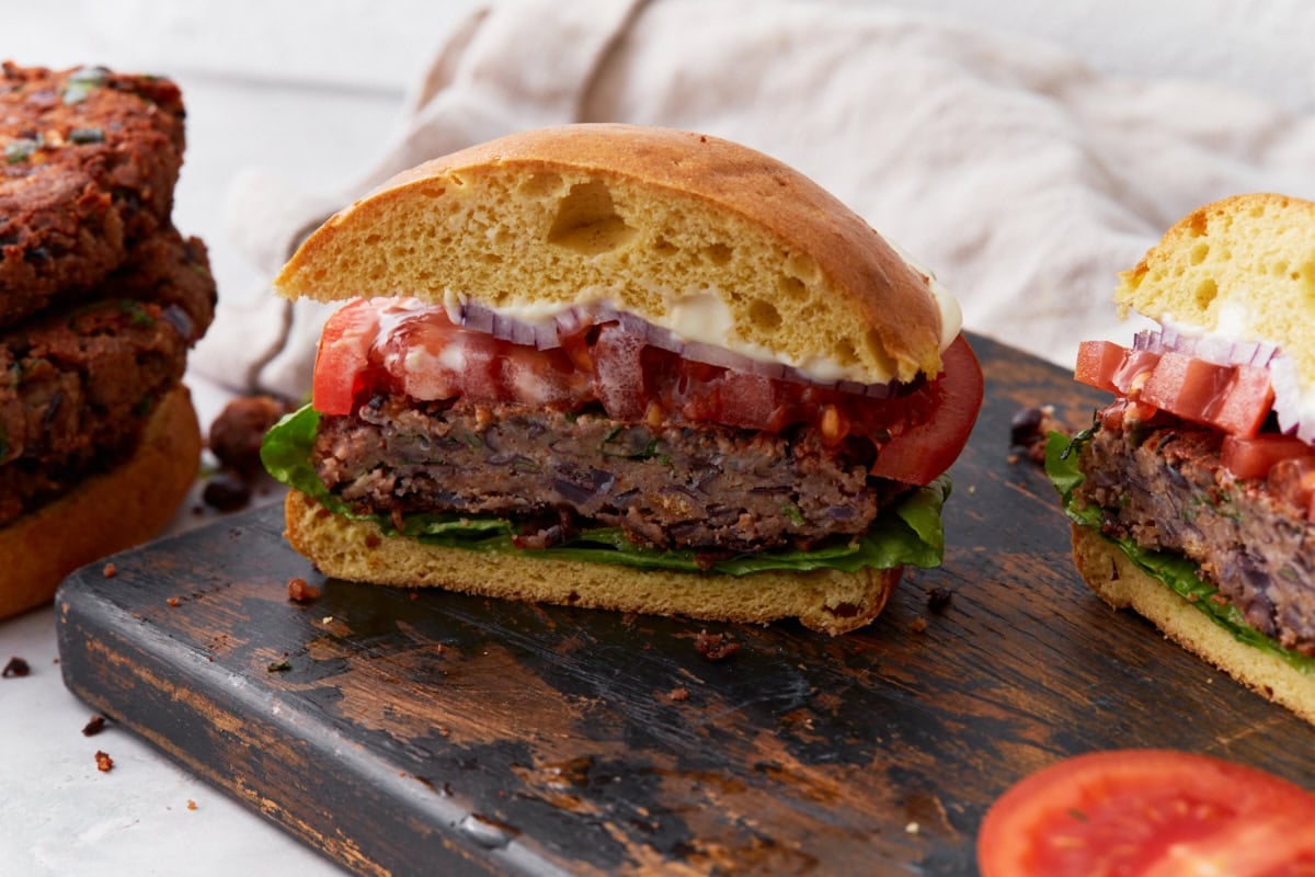 A close-up of a gluten-free black bean burger sliced in half and stacked with lettuce, tomato, red onion, and mayo on a toasted gluten-free bun.