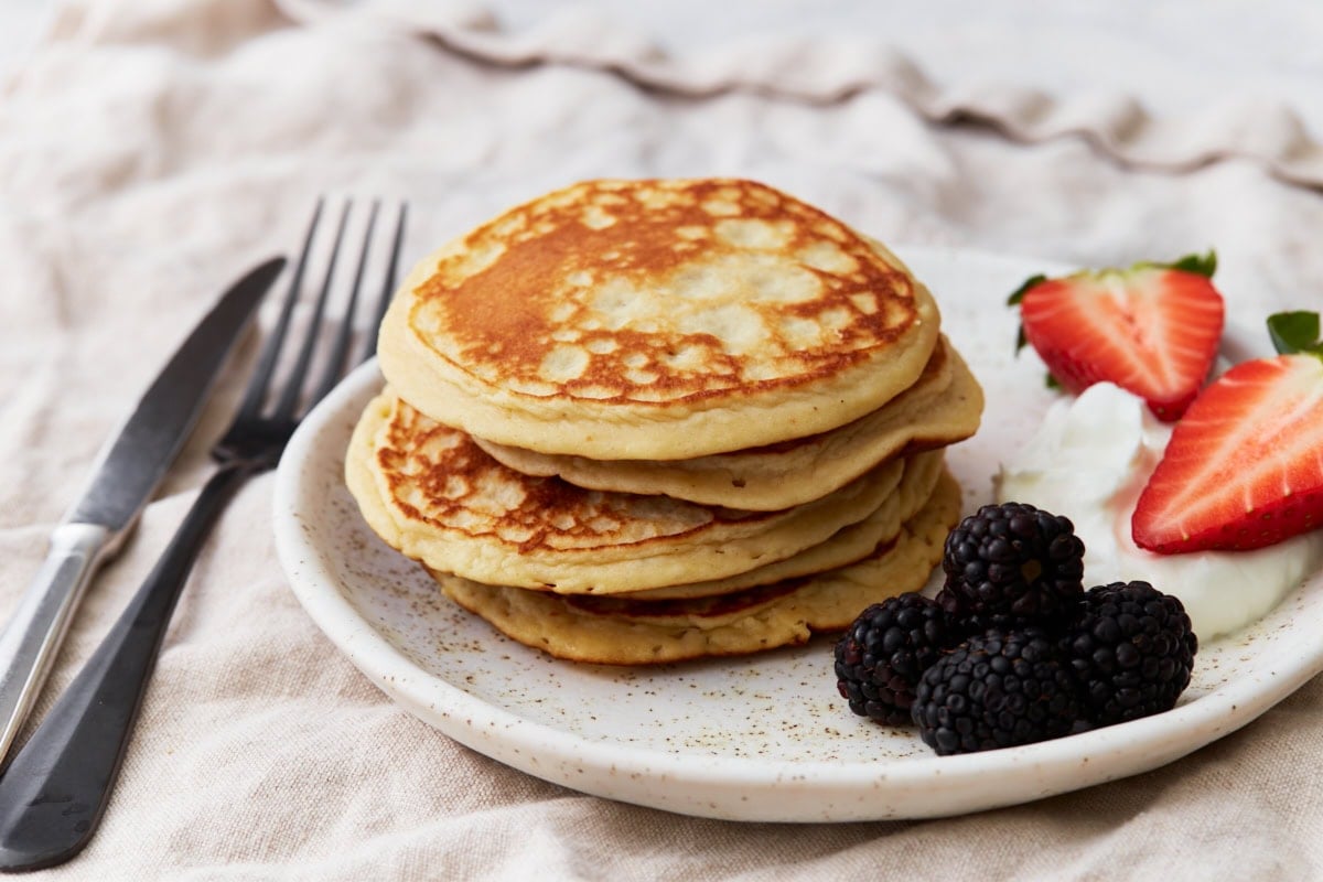 A close-up of a stack of gluten-free protein pancakes on a white plate with berries and Greek yogurt, styled for serving.