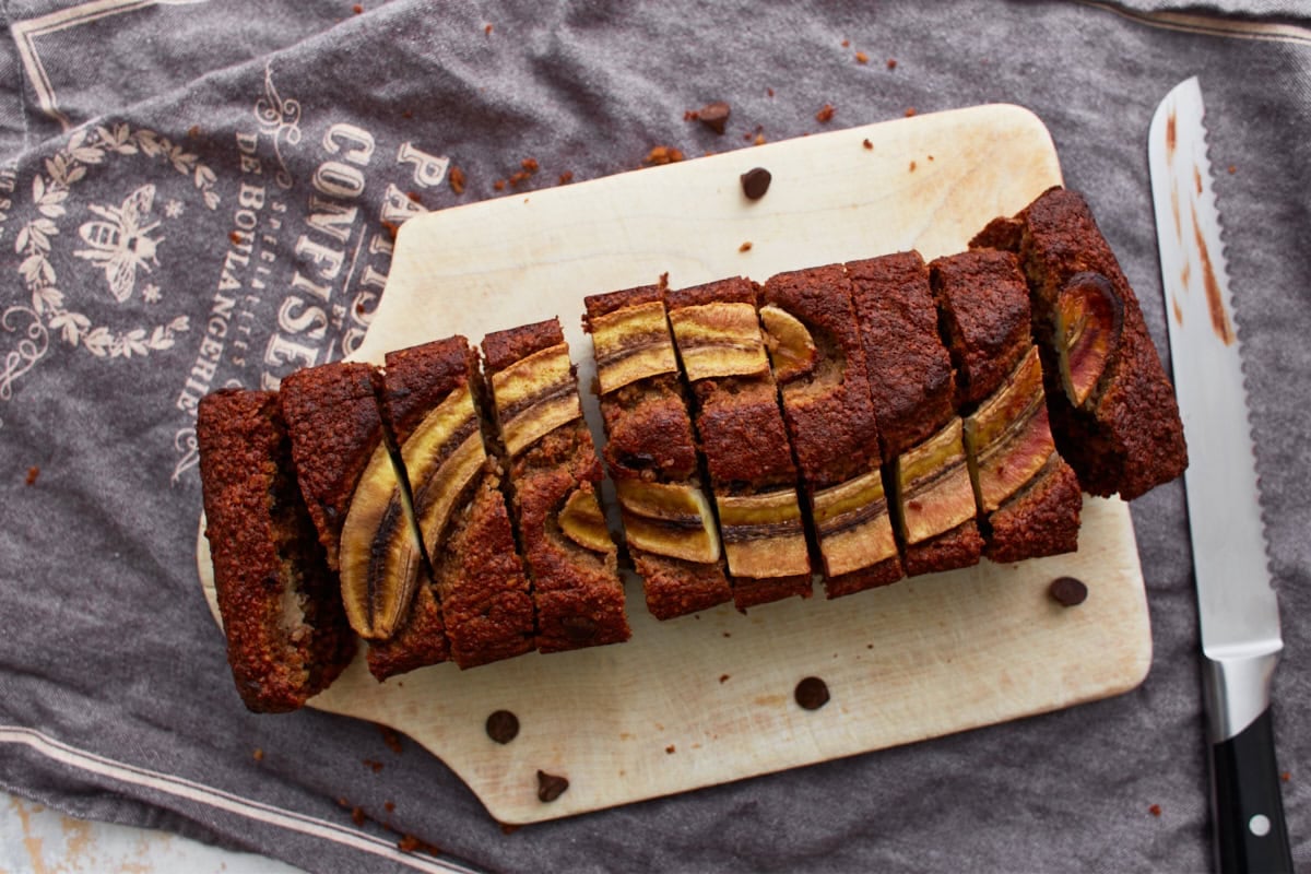 A top-down view of a fully sliced loaf of banana oatmeal bread on a wooden cutting board, with a serrated knife nearby.
