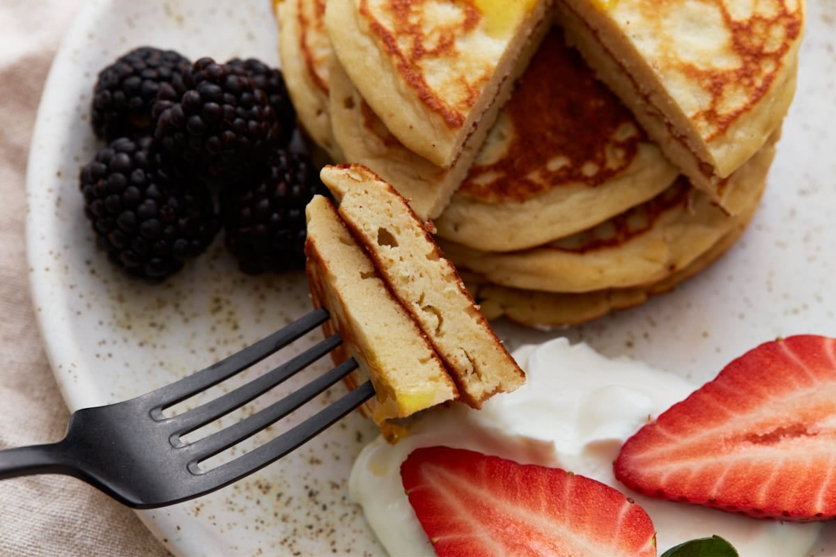 A fork holding a bite of fluffy pancake in front of a stack, with Greek yogurt, sliced strawberries, and blackberries in the background.