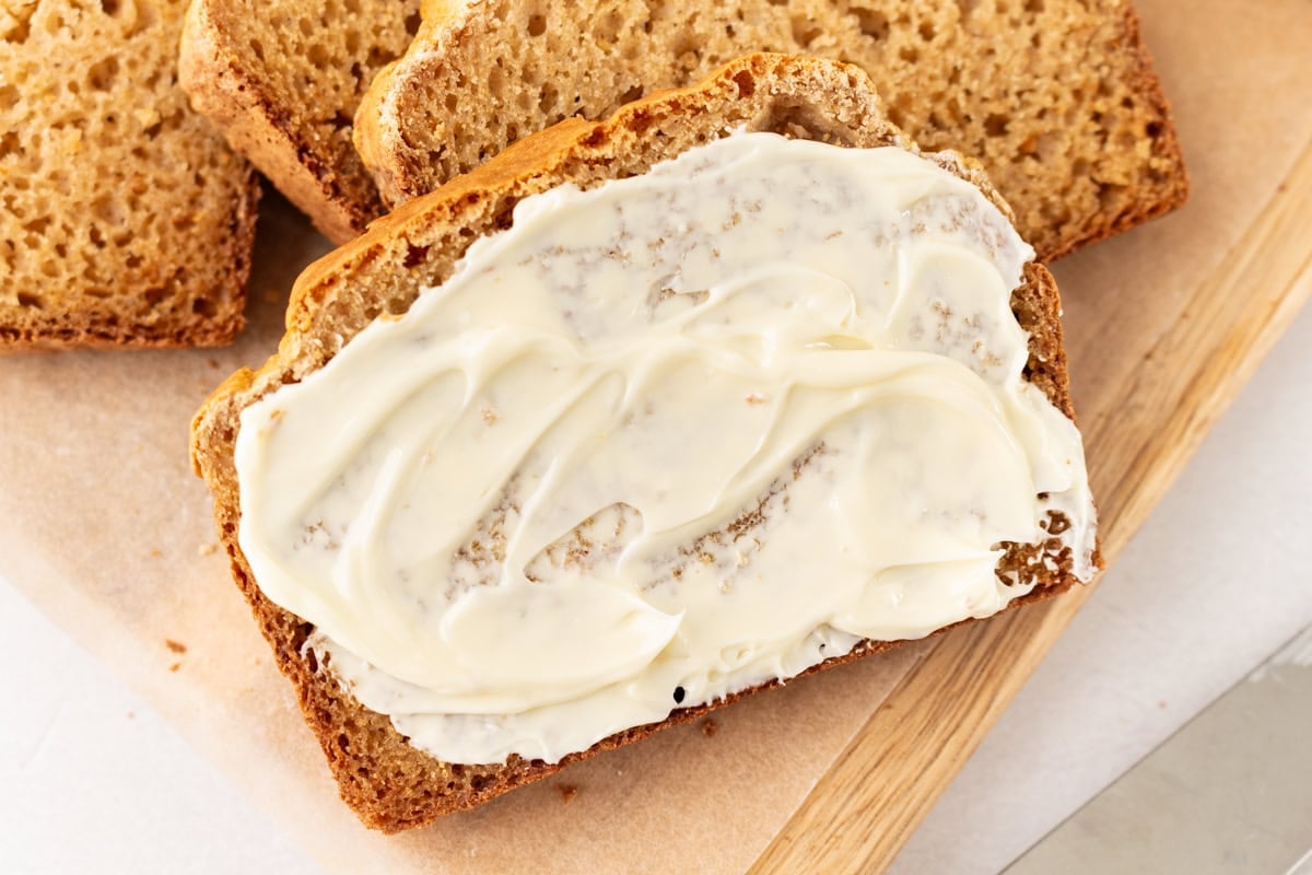 Close-up of a slice of cassava bread generously spread with butter, surrounded by other slices on parchment paper.