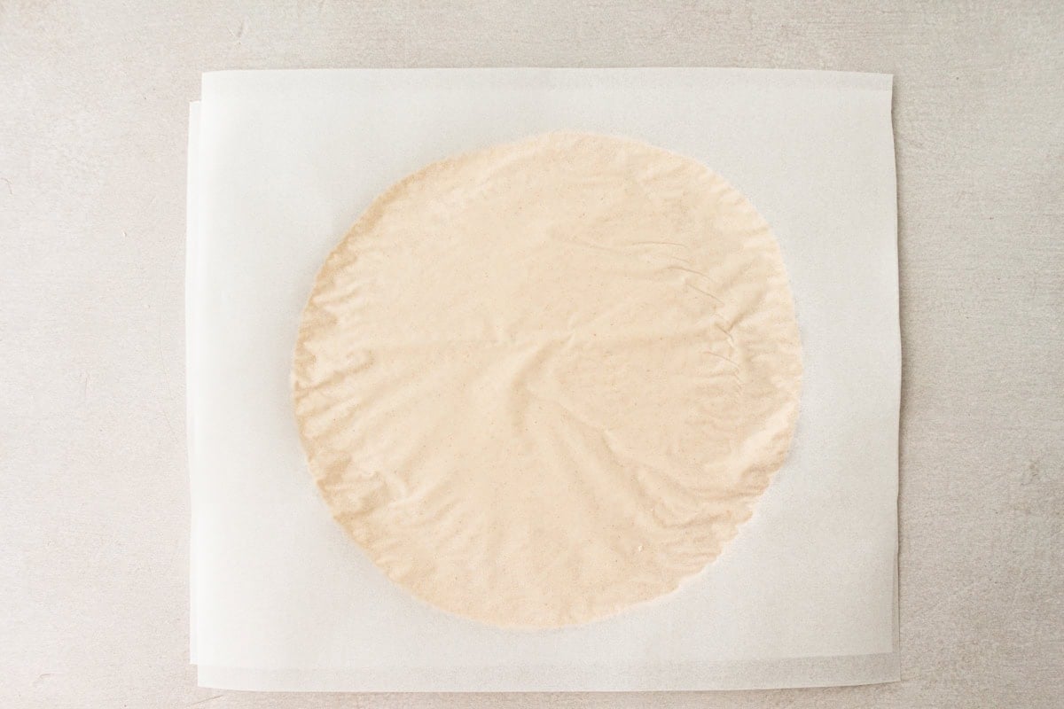 A rolled-out cassava tortilla between two sheets of parchment paper, ready to cook.