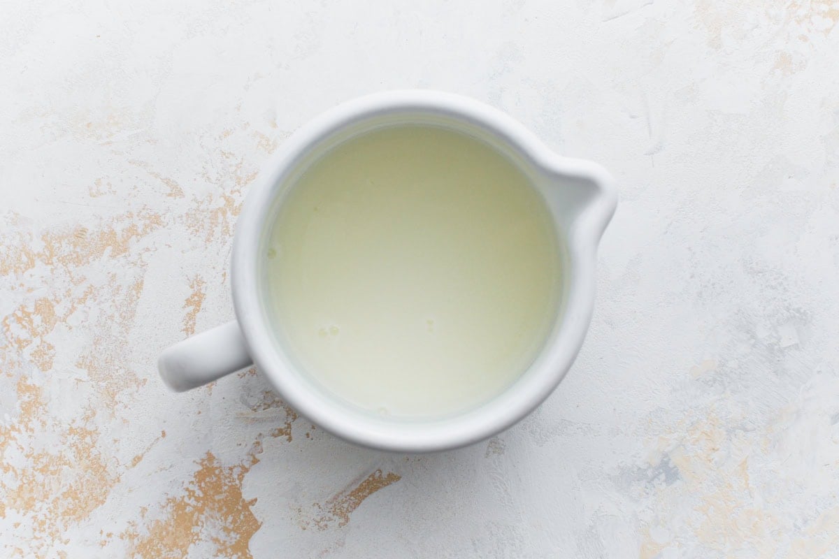 Top-down view of a small white ceramic pitcher filled with buttermilk, set on a light textured background.