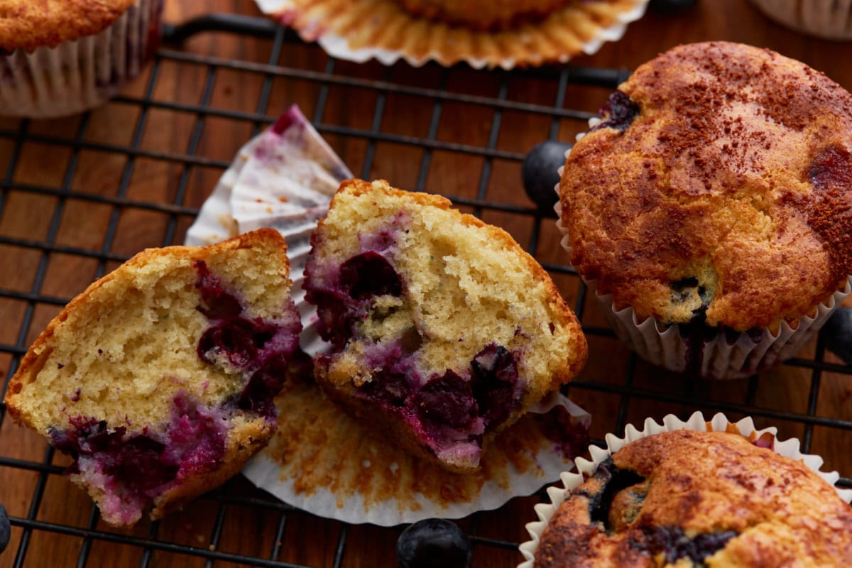 One gluten-free blueberry muffin cut in half to reveal its soft, fluffy crumb and bursting blueberries, surrounded by whole muffins on a cooling rack.