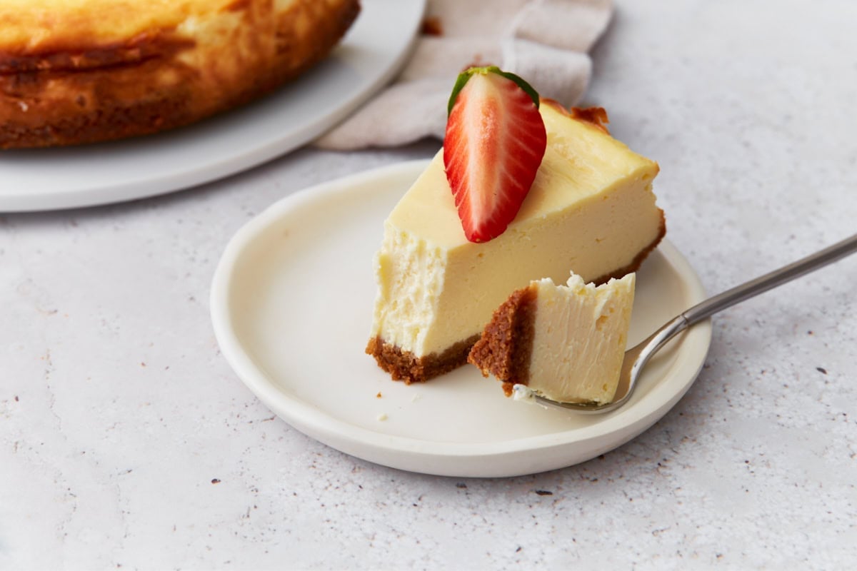 Slice of gluten-free cheesecake with a bite taken out, topped with a strawberry, sitting on a small white plate with a fork.