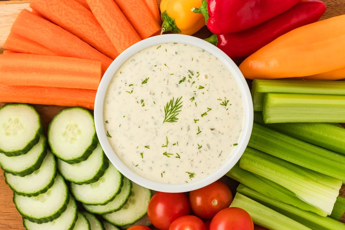 Bowl of dairy-free ranch dressing garnished with dill, surrounded by fresh sliced cucumbers, carrots, celery, and colorful mini bell peppers on a wooden board.