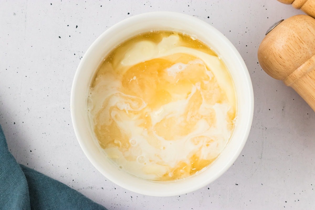 Overhead view of a white bowl with mayonnaise and vinegar beginning to mix together on a speckled white surface.