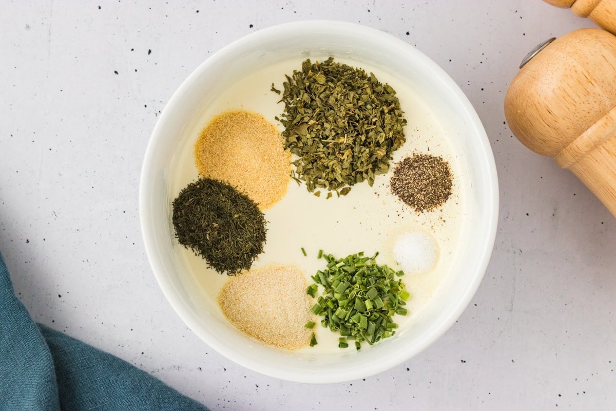 Overhead view of a bowl filled with dairy-free ranch base topped with piles of dried herbs and spices, ready to be mixed.