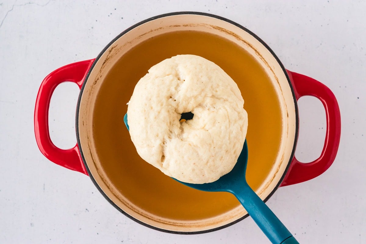 A raw bagel being lifted from a pot of boiling water with honey, shown mid-boil in a red Dutch oven.