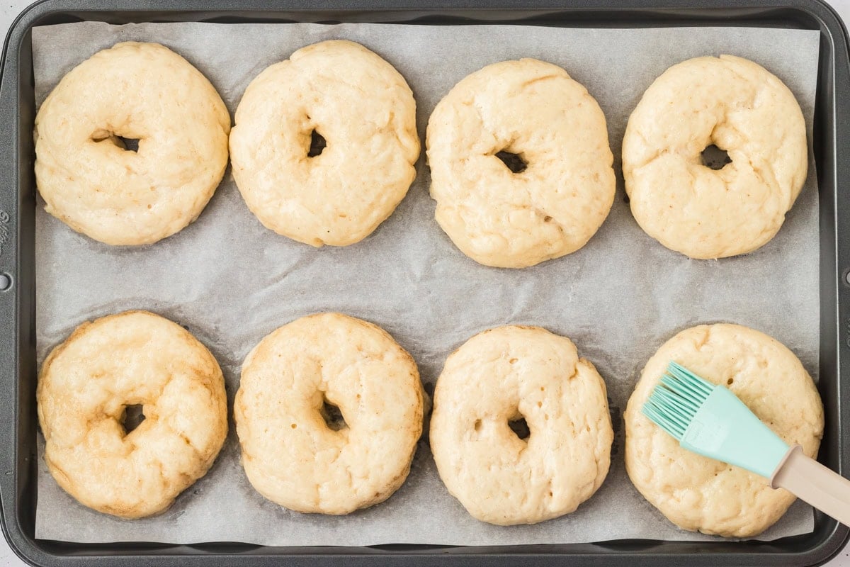 Par-boiled gluten-free bagels on a baking tray being brushed with egg wash before baking.