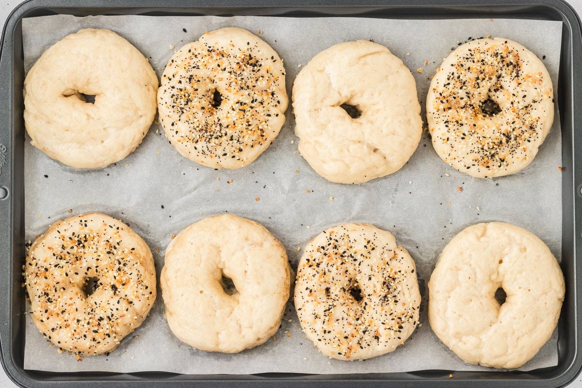 Unbaked gluten-free bagels on a parchment-lined baking sheet, some topped with everything seasoning and others plain, ready to go in the oven.