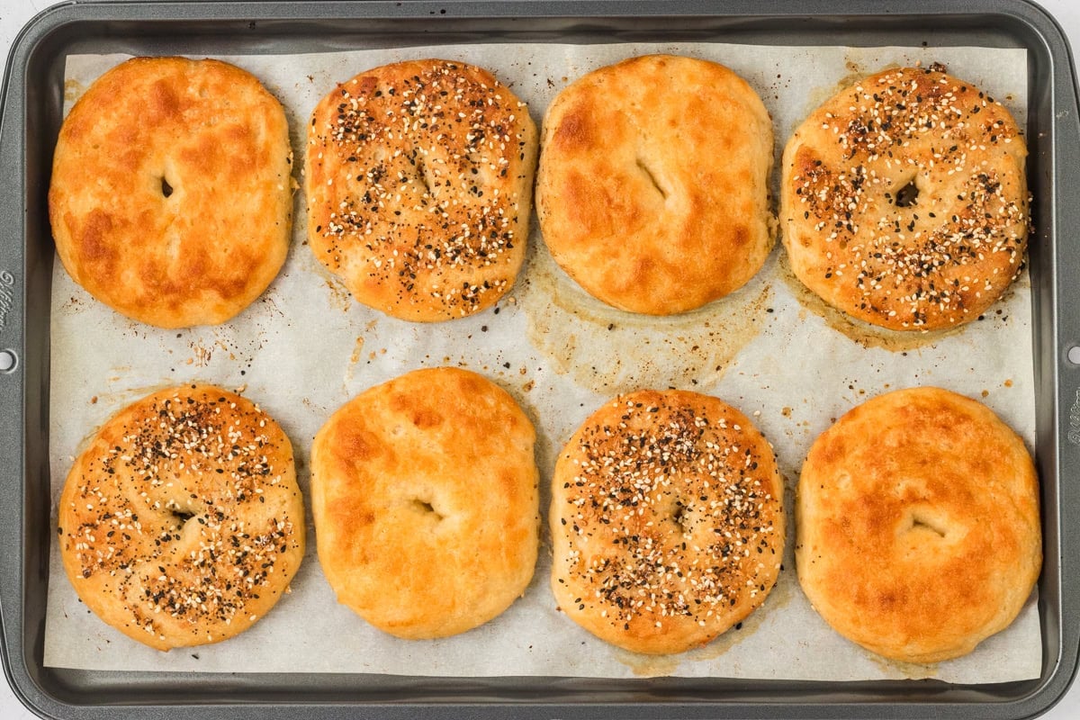 Golden brown gluten-free bagels just out of the oven on a baking sheet, with a mix of plain and everything bagel toppings.