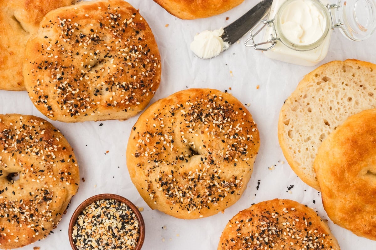 Overhead shot of freshly baked gluten-free bagels on parchment paper, with a dish of everything bagel seasoning, a jar of cream cheese, and one bagel sliced open to show the soft crumb inside.