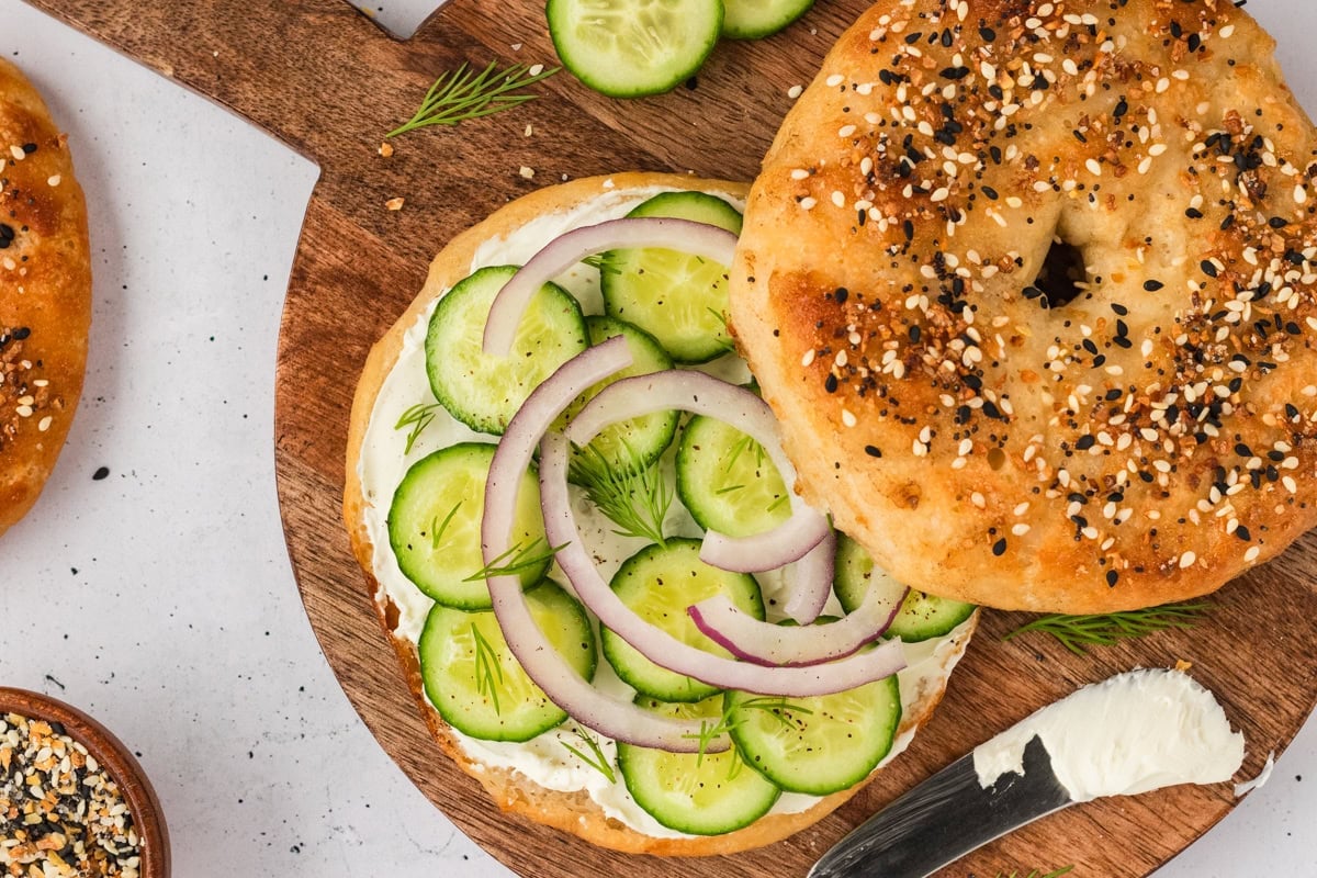 A gluten-free everything bagel sandwich on a wooden board, filled with cream cheese, cucumber slices, red onion, and fresh dill, with a knife and extra cream cheese nearby.
