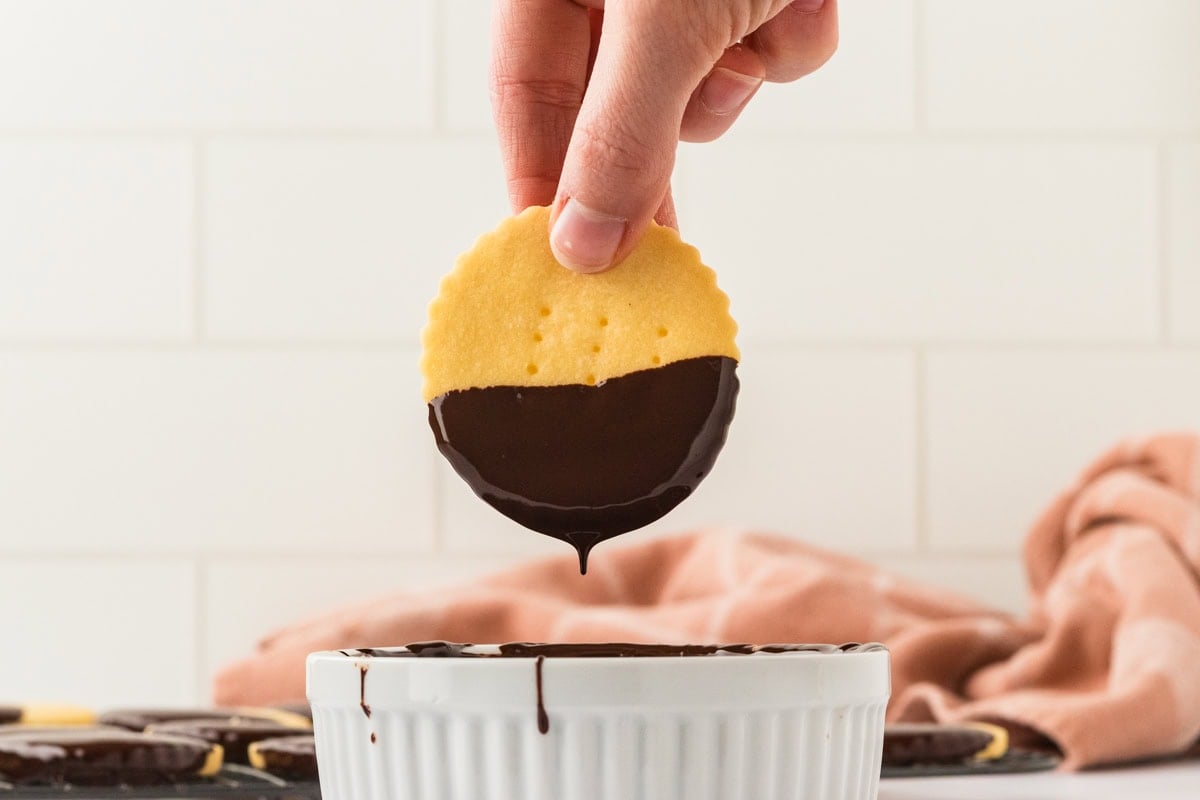 A hand holding a shortbread cookie halfway dipped in melted chocolate above the bowl.