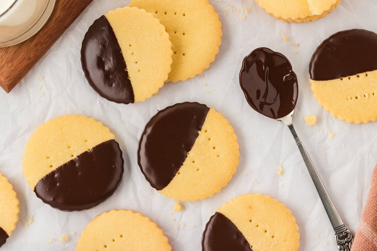 Several shortbread cookies laid out on parchment paper, half-dipped in chocolate, with a spoon of melted chocolate nearby.