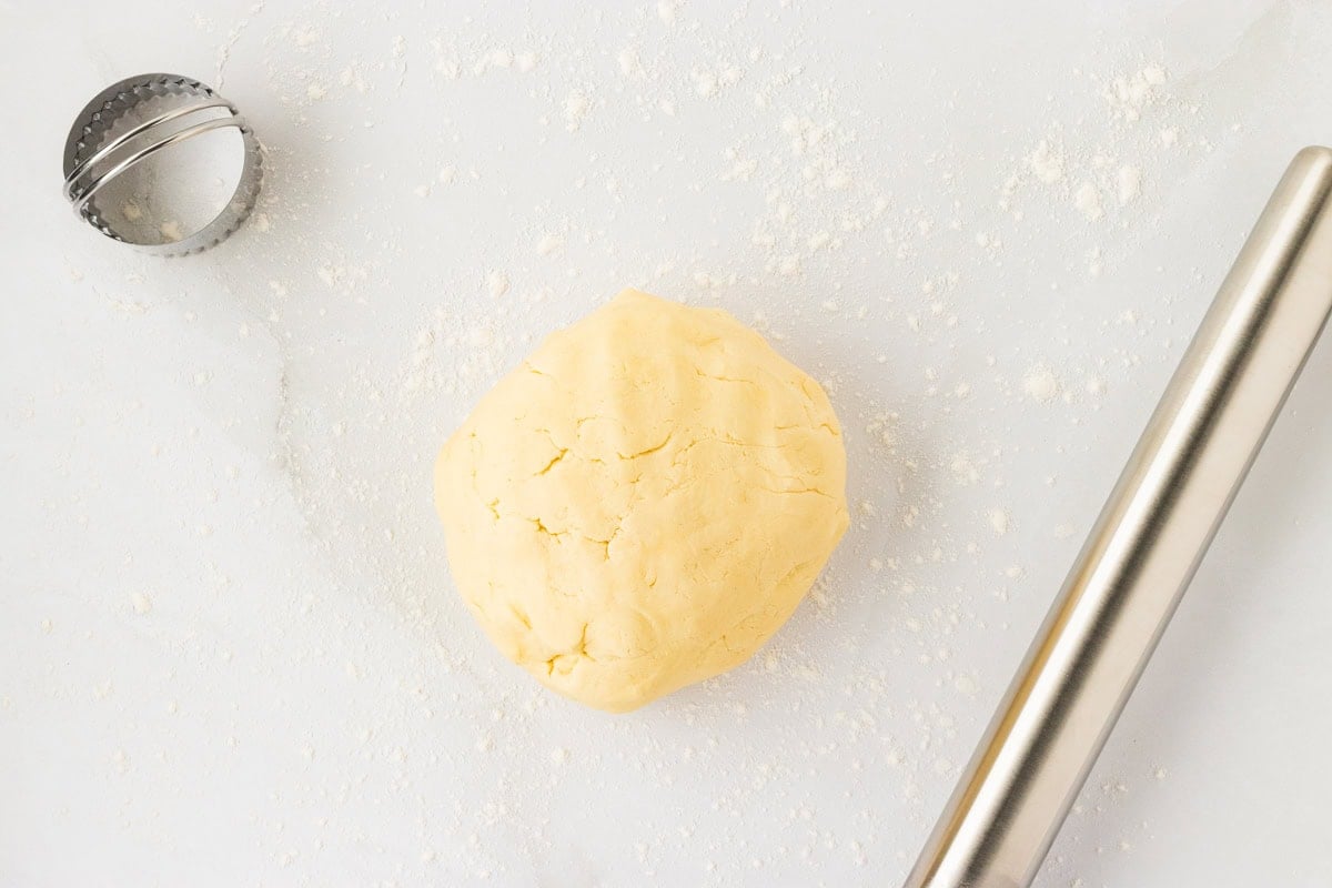 Ball of gluten-free shortbread cookie dough placed on a lightly floured surface with a cookie cutter and rolling pin nearby.