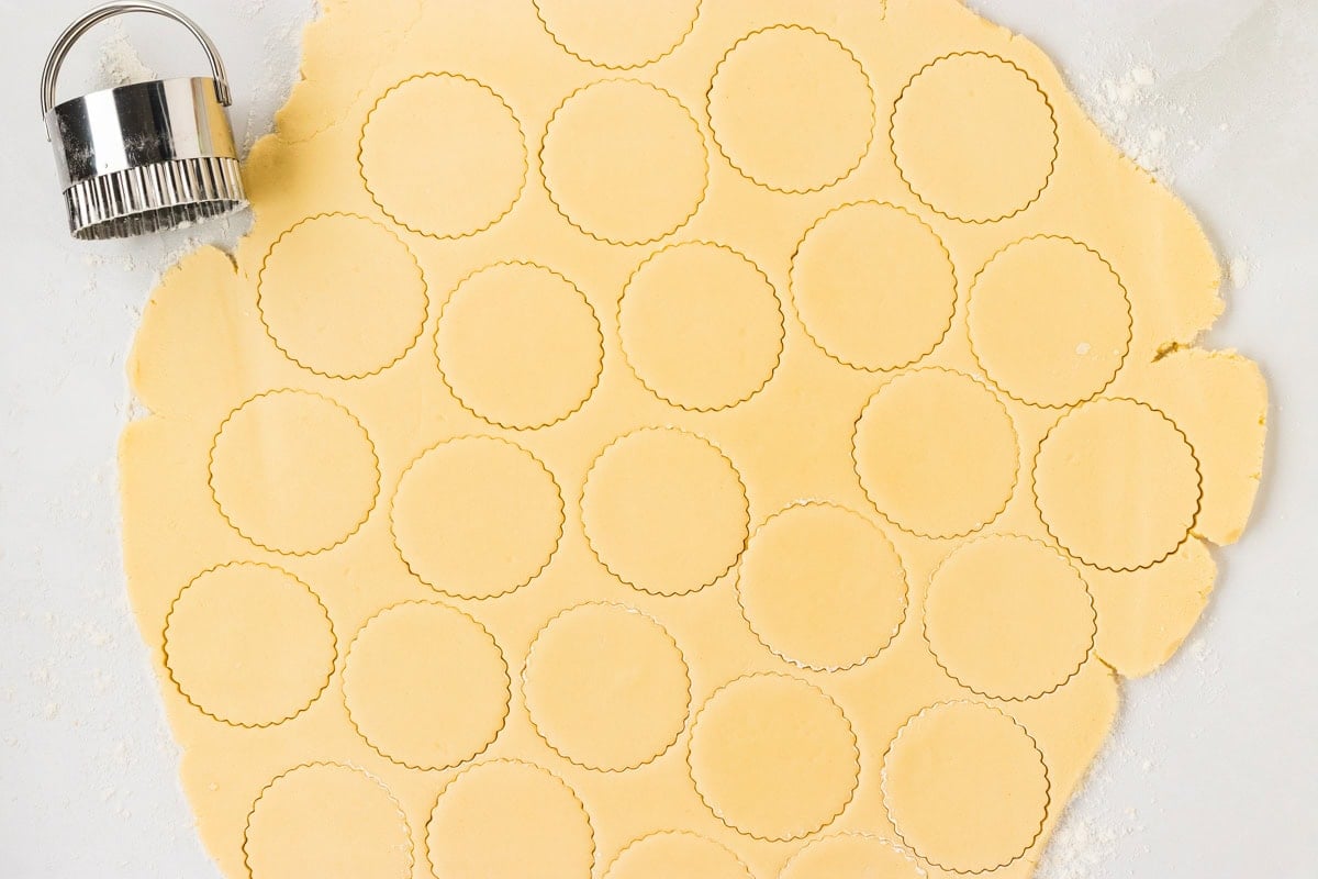 Rolled out dough with round scalloped cookie shapes cut out, ready to be transferred to the baking sheet.