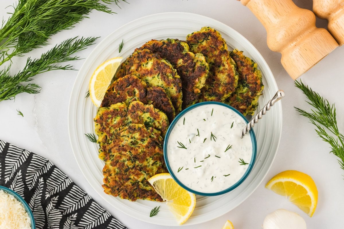 A serving platter of pan-fried zucchini fritters arranged around a bowl of creamy dill dipping sauce, garnished with fresh dill and lemon wedges.