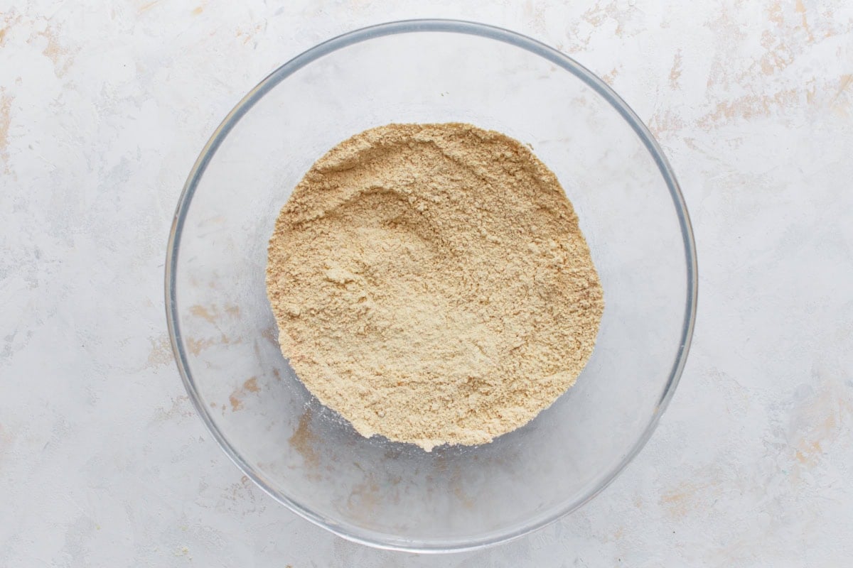 A glass mixing bowl filled with a light yellow blend of dry ingredients, including chickpea flour and spices.