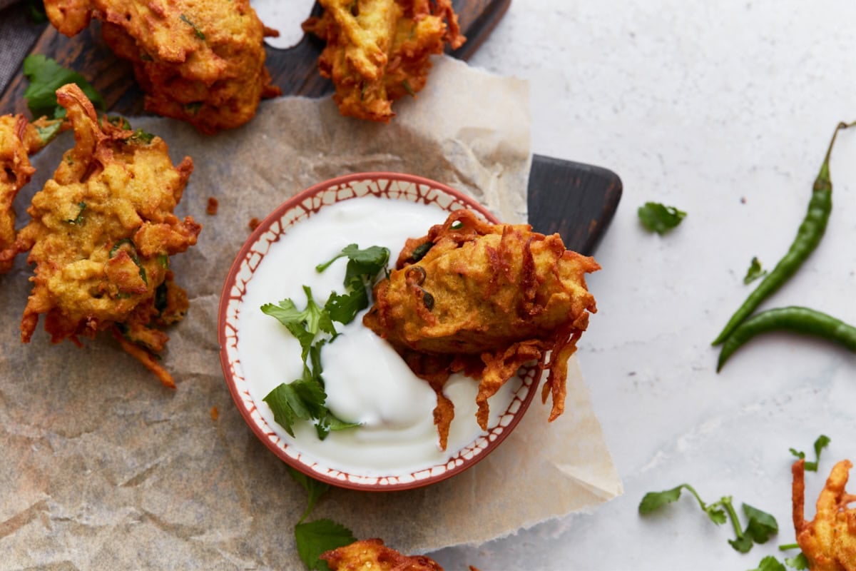 A single pakora dipped into a small bowl of yogurt sauce, garnished with chopped cilantro for serving.