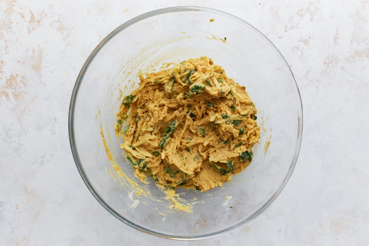 Pakora batter in a bowl with shredded potatoes, spinach, and cilantro fully mixed into the thick spiced paste.