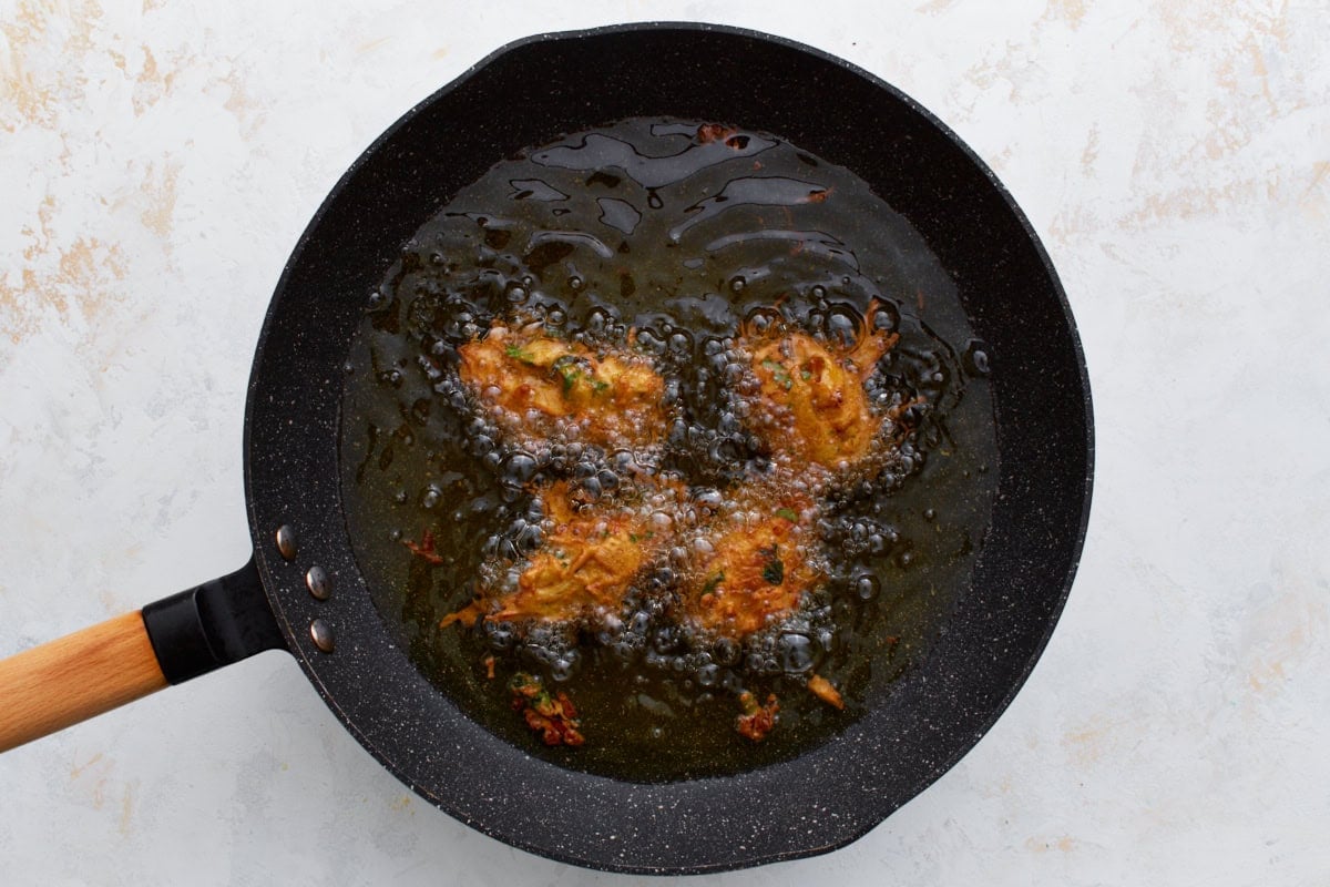 Several pakoras sizzling in hot oil in a black frying pan, bubbling as they turn golden and crisp.