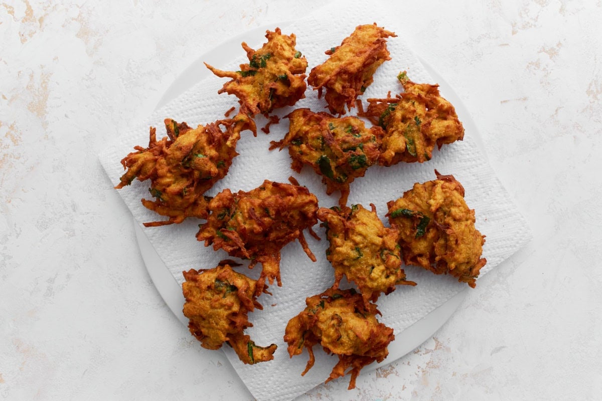 Freshly fried pakoras cooling on a white paper towel-lined plate, showing their crispy, golden texture.