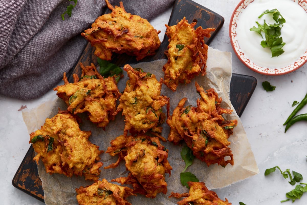 A styled shot of crispy pakoras served on parchment paper with a side of creamy dipping sauce and fresh green chili peppers scattered around.