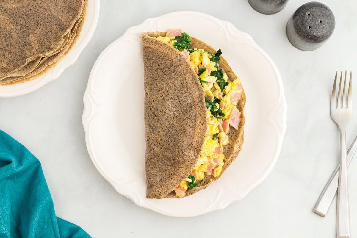 A buckwheat crepe folded in half and filled with scrambled eggs, ham, and sautéed spinach on a white plate, with salt and pepper shakers and utensils nearby.