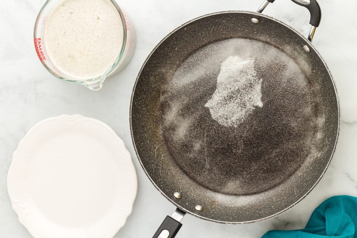 A non-stick frying pan sprayed with cooking oil, sitting next to a measuring cup of crepe batter and an empty white plate.