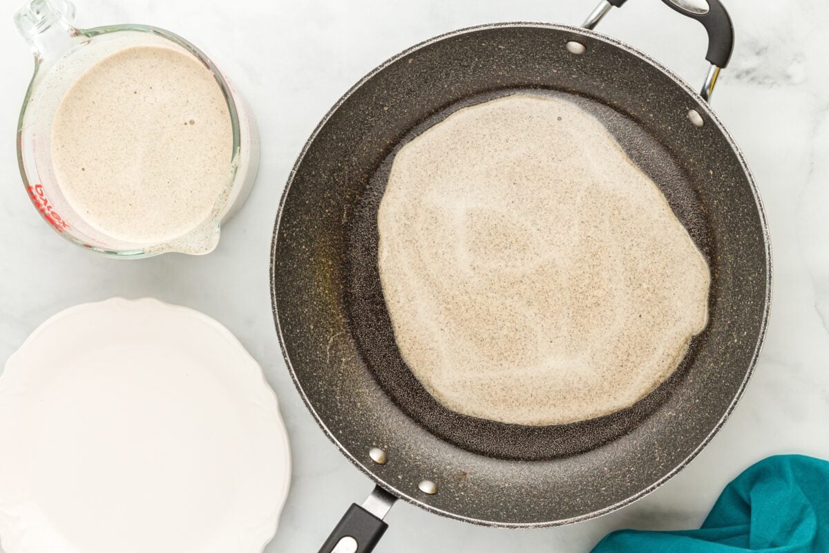 A buckwheat crepe cooking in a skillet, forming a thin circular layer, with crepe batter in a measuring cup nearby.
