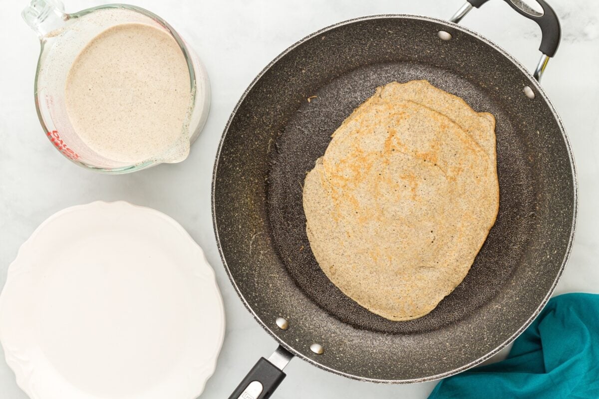 A golden-brown buckwheat crepe cooked on one side in a skillet, with a measuring cup of batter and an empty plate beside it.