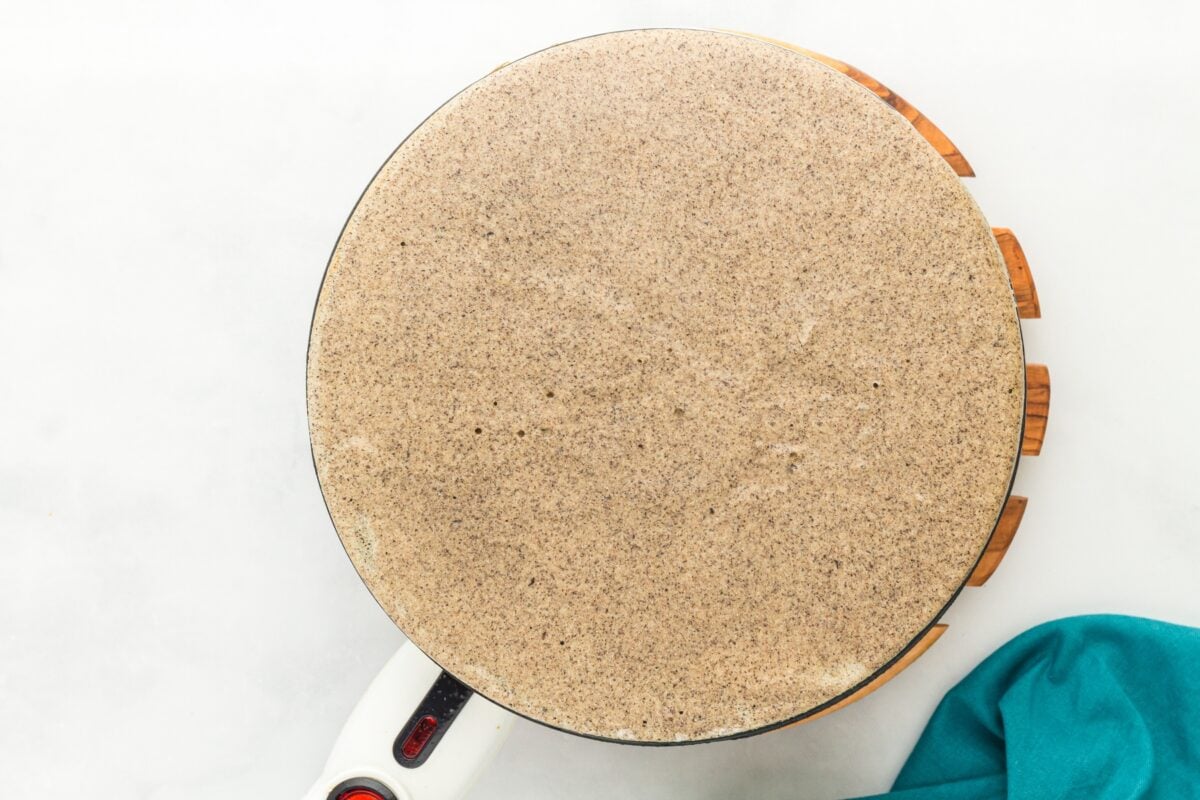 A thin buckwheat crepe cooking on an electric crepe maker with a smooth, round surface.