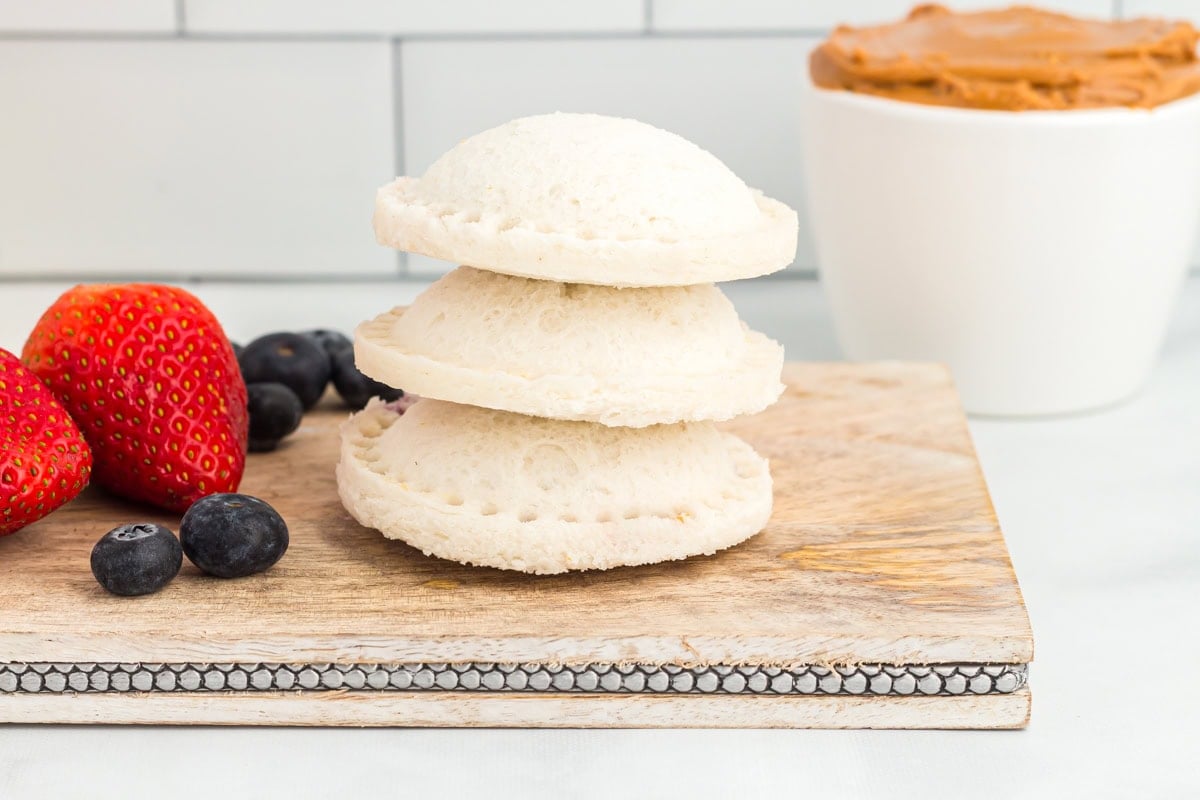A stack of three homemade gluten-free uncrustables on a rustic wooden board with fresh strawberries and blueberries in the background.