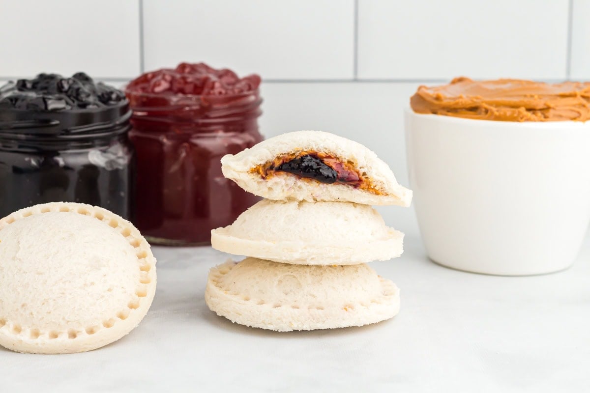 A close-up of a stack of homemade gluten-free uncrustables, with one cut open to show the peanut butter and jam filling, jars of jam and a bowl of peanut butter in the background.