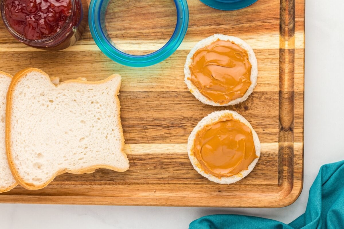 Two gluten-free bread circles spread with a layer of peanut butter on a wooden cutting board, next to more bread slices and jars of jam.