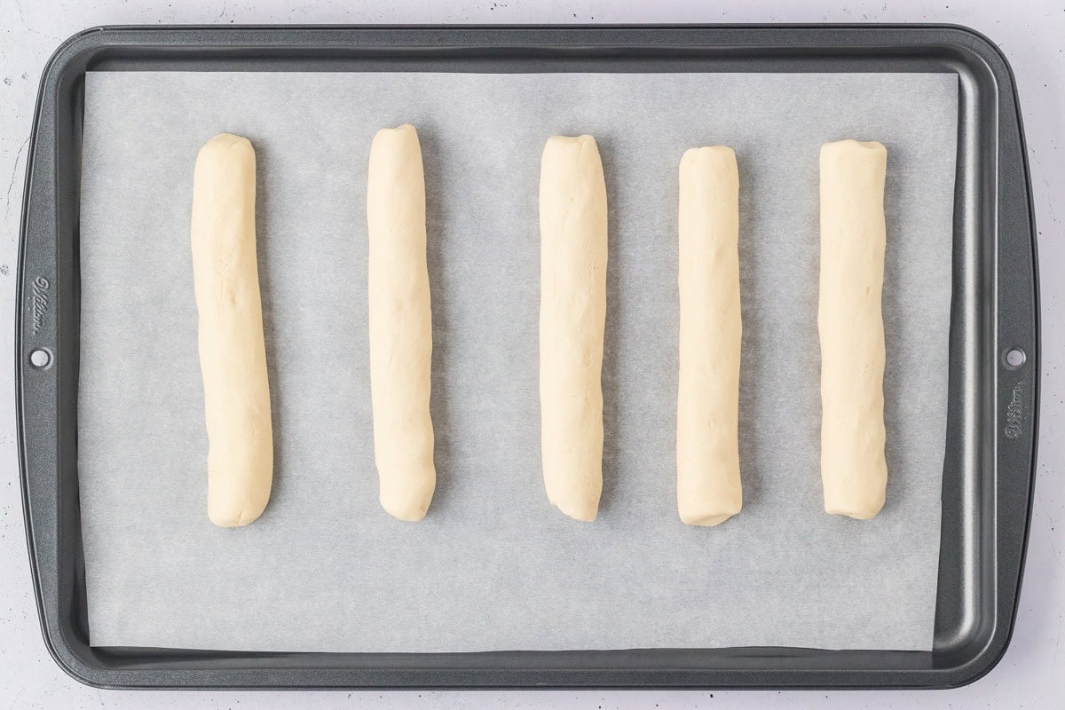 Five shaped gluten-free breadsticks evenly spaced on a parchment-lined baking sheet.