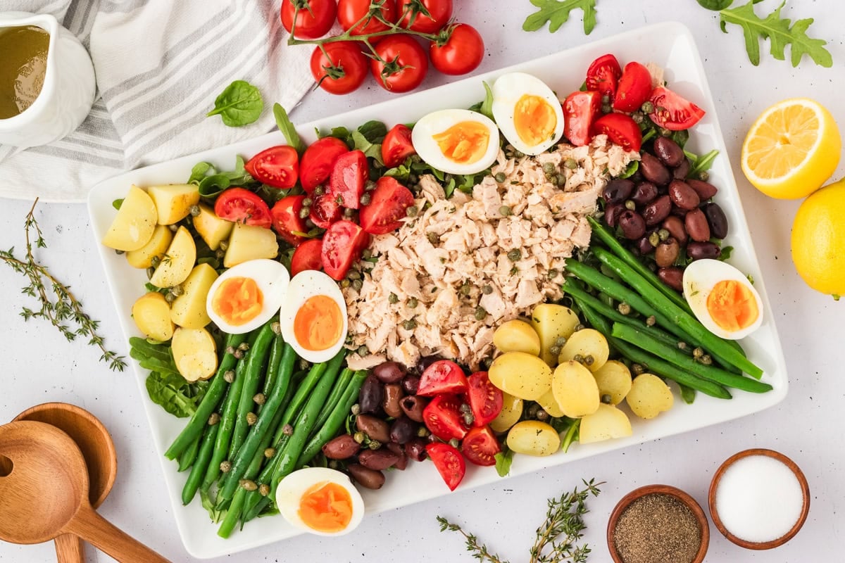 Overhead view of the completed Tuna Nicoise Salad, styled with fresh thyme, lemons, tomatoes, and dressing on the side.